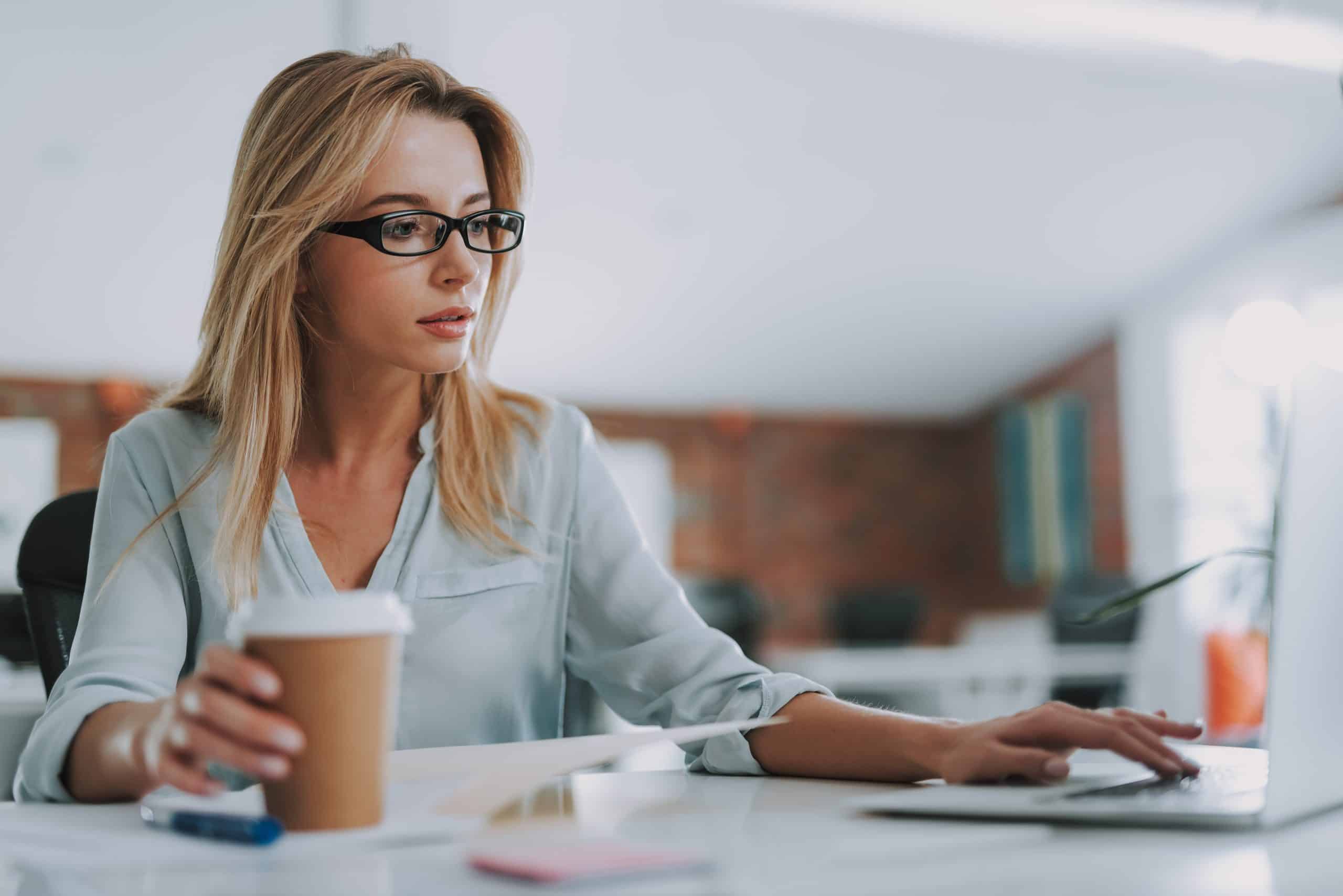 Thoughtful woman touching the keyboard of her laptop Close up photo of the office worker sitting with a carton cup of coffee and looking attentively at the laptop while touching it